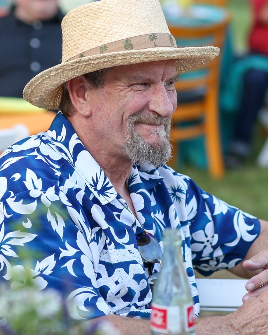 Tim in a straw hat and a blue Hawaiian shirt, smiling at an outdoor gathering.